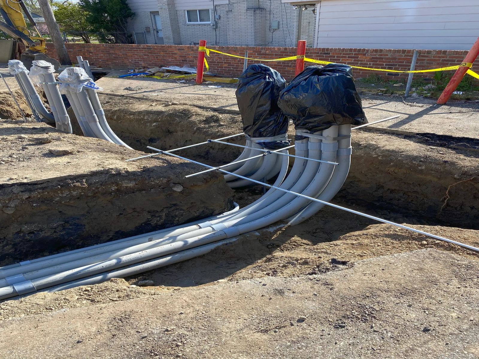 A bundle of grey conduits curving up out of an open trench beside a brick wall, taped off with caution ribbon.