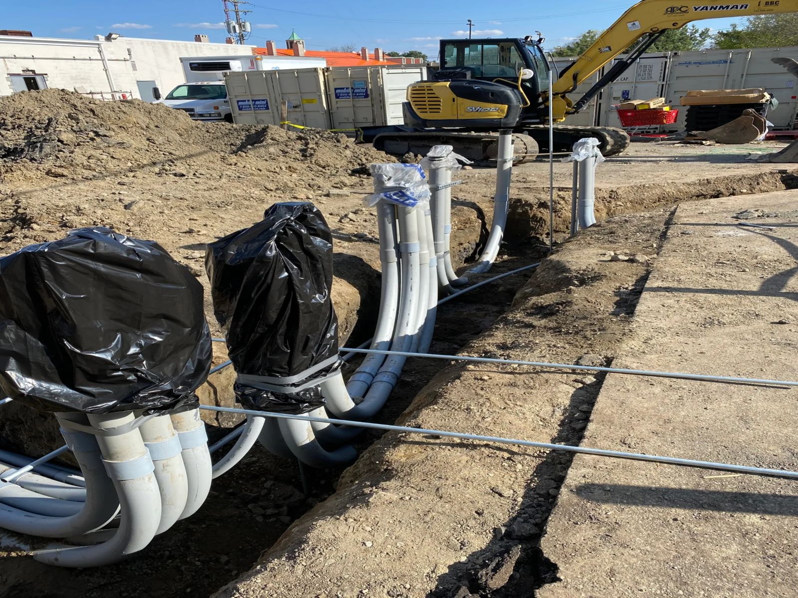A bank of underground conduits sweeping up out of a trench, with an excavator parked behind it.