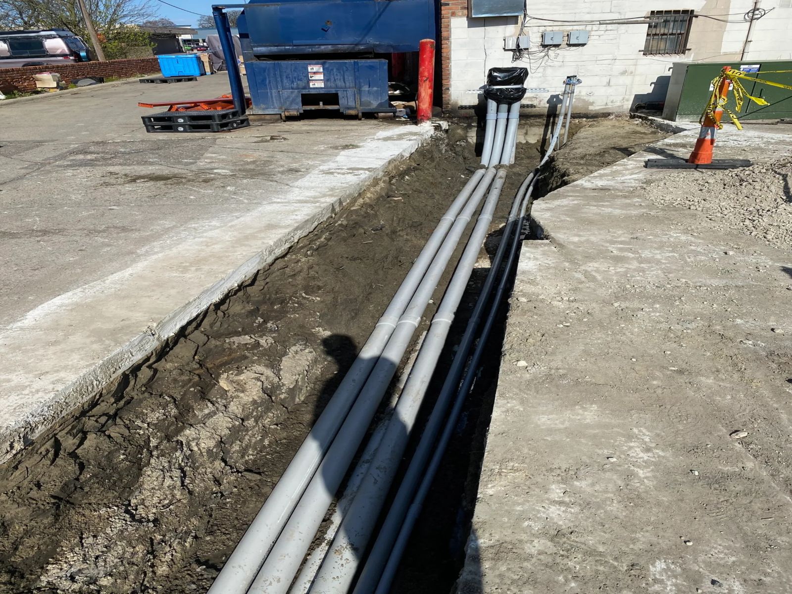 A long trench cut across a commercial lot with grey conduit and cable pulled the length of it.